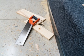 On a speckled concrete floor, a handsaw with an orange and black handle is placed on three pieces of light-colored wooden plank alongside a black carpeted edge.