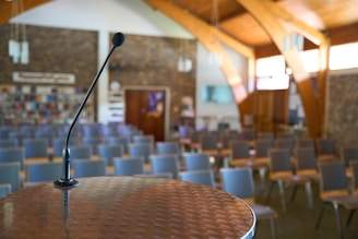 A microphone is positioned on a podium in an empty auditorium, with rows of chairs facing it. The room features wooden beams, a brick wall at the back with bookshelves, and several pendant lights hanging from the ceiling.