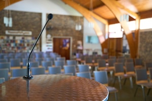 A microphone is positioned on a podium in an empty auditorium, with rows of chairs facing it. The room features wooden beams, a brick wall at the back with bookshelves, and several pendant lights hanging from the ceiling.