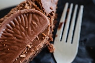 A close-up of a chocolate dessert featuring a textured chocolate piece on top and a silver fork placed beside it. The dessert has a rich, crumbly appearance and multiple layers coated with chocolate, set against a dark background.