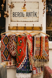Colorful, patterned rugs hang on wooden hangers beneath a sign that reads 'BEROL ANTIK.' The sign is mounted on a textured white wall, and dry leaves dangle from above, adding a rustic touch to the scene.