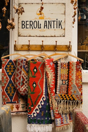 Colorful, patterned rugs hang on wooden hangers beneath a sign that reads 'BEROL ANTIK.' The sign is mounted on a textured white wall, and dry leaves dangle from above, adding a rustic touch to the scene.