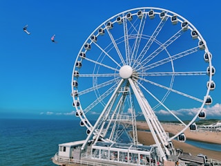 A large Ferris wheel is situated near a beach, with a view of the ocean in the background. Two people are zip lining in the sky, adding a sense of adventure to the scene. The sky is clear and blue, and the beach has patches of greenery with people visible along the shoreline.