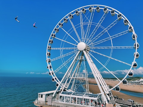A large Ferris wheel is situated near a beach, with a view of the ocean in the background. Two people are zip lining in the sky, adding a sense of adventure to the scene. The sky is clear and blue, and the beach has patches of greenery with people visible along the shoreline.