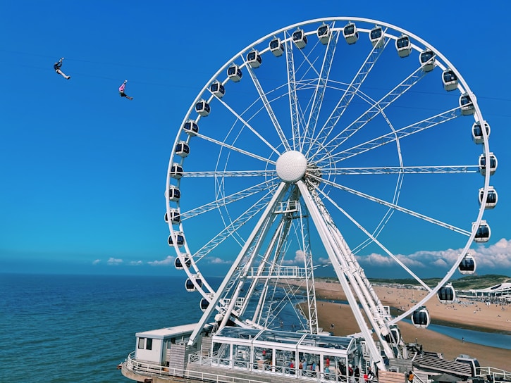 A large Ferris wheel is situated near a beach, with a view of the ocean in the background. Two people are zip lining in the sky, adding a sense of adventure to the scene. The sky is clear and blue, and the beach has patches of greenery with people visible along the shoreline.