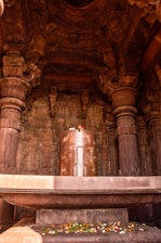 A large stone structure resembling a Shiva Lingam is situated in the center of a temple's interior. The structure is adorned with floral decorations, and the surrounding architecture features intricate carvings and columns. Ambient lighting highlights the texture and detail of the stonework.