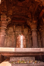 A large stone structure resembling a Shiva Lingam is situated in the center of a temple's interior. The structure is adorned with floral decorations, and the surrounding architecture features intricate carvings and columns. Ambient lighting highlights the texture and detail of the stonework.
