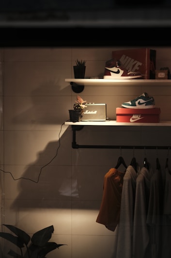 A stylish shelf display illuminated with warm lighting includes a pair of sneakers on shoe boxes, a small plant, and a speaker. Hanging clothes are visible below the shelf in muted tones, with some greenery at the bottom left corner.
