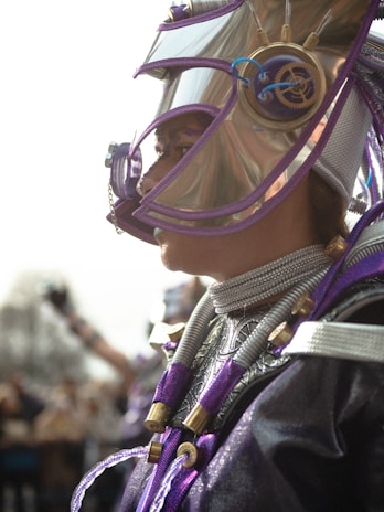 A person wearing an elaborate and futuristic costume, featuring metallic elements and intricate details. The headgear includes a helmet with a reflective visor and elaborate mechanical embellishments, predominantly in purple and silver tones. The outfit is adorned with textured patterns and cord-like attachments, enhancing the overall sci-fi appearance.