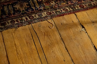 Worn, patterned rug partially covering a rustic wooden floor with visible grains and gaps between the planks. The rug displays intricate designs with floral and geometric motifs in earthy tones.