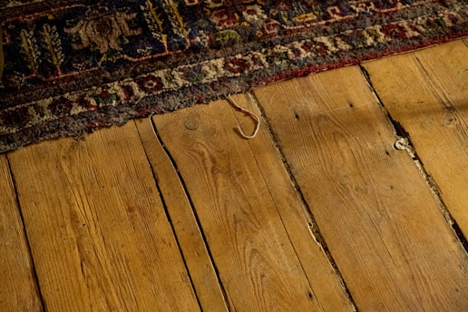 Worn, patterned rug partially covering a rustic wooden floor with visible grains and gaps between the planks. The rug displays intricate designs with floral and geometric motifs in earthy tones.