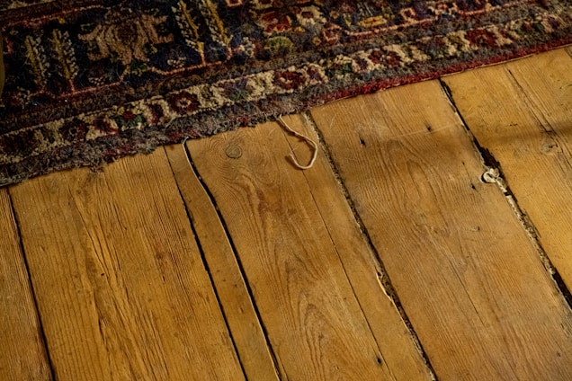Worn, patterned rug partially covering a rustic wooden floor with visible grains and gaps between the planks. The rug displays intricate designs with floral and geometric motifs in earthy tones.