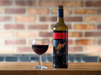 A bottle of wine labeled Yellow Tail Cabernet Sauvignon stands on a wooden surface next to a filled wine glass. The background features a blurred brick wall, enhancing the warm atmosphere.