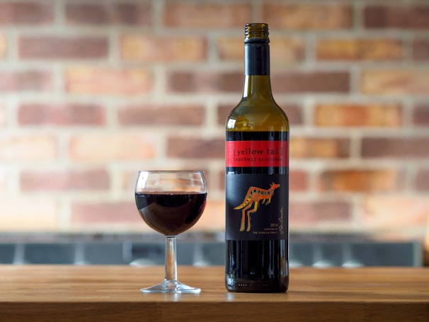 A bottle of wine labeled Yellow Tail Cabernet Sauvignon stands on a wooden surface next to a filled wine glass. The background features a blurred brick wall, enhancing the warm atmosphere.