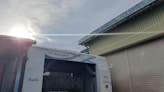 A car wash facility is shown with a blue and white sign reading 'Beauty Wash'. The facility is positioned between two buildings with corrugated metal shutters. The sun is partially visible in the sky, casting a bright flare across the upper part of the image. Snow or frost is visible on the rooftops and the car wash structure.