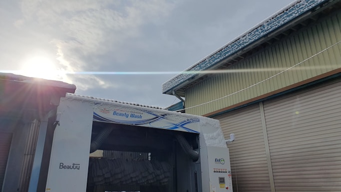 A car wash facility is shown with a blue and white sign reading 'Beauty Wash'. The facility is positioned between two buildings with corrugated metal shutters. The sun is partially visible in the sky, casting a bright flare across the upper part of the image. Snow or frost is visible on the rooftops and the car wash structure.