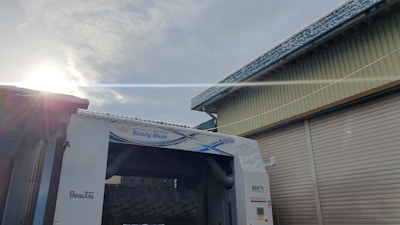 A car wash facility is shown with a blue and white sign reading 'Beauty Wash'. The facility is positioned between two buildings with corrugated metal shutters. The sun is partially visible in the sky, casting a bright flare across the upper part of the image. Snow or frost is visible on the rooftops and the car wash structure.