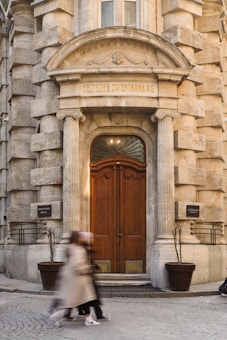 An ornate entrance of an old stone building with a prominent wooden door framed by classical columns and an arch. The inscription 'DEUTSCHE ORIENTBANK A.G.' is visible above the door. Two blurred figures appear to be walking past the entrance on the cobblestone street in front.