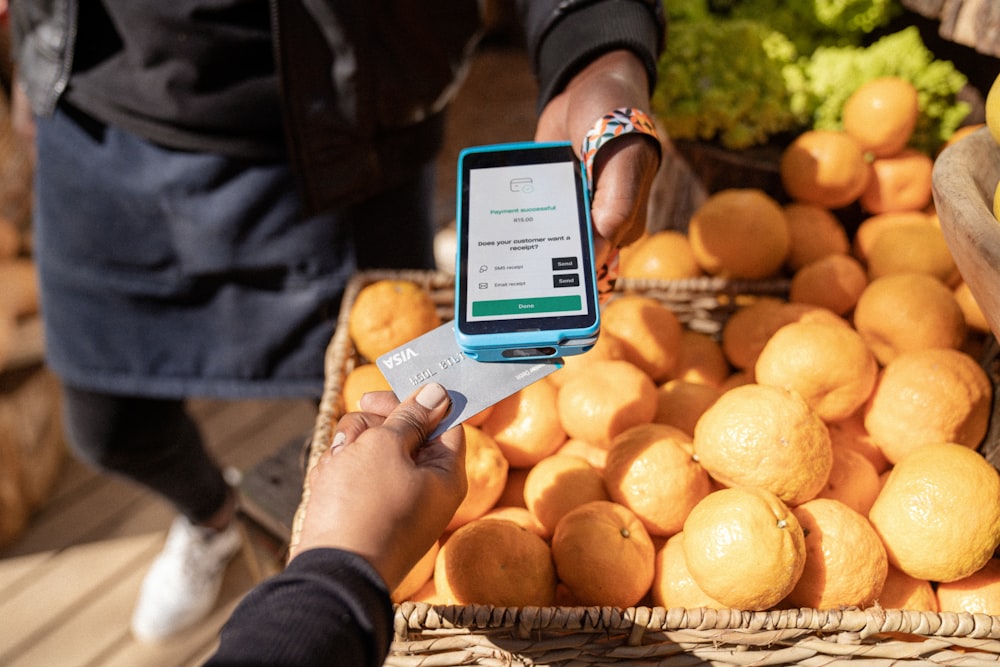 a person holding a smart phone in front of a basket of oranges