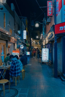 A narrow, dimly lit urban alleyway features several small eateries and shops with bright signboards written in Japanese. People are sitting on stools outside, enjoying meals and drinks. The atmosphere is lively and bustling, typical of a city nightlife scene.