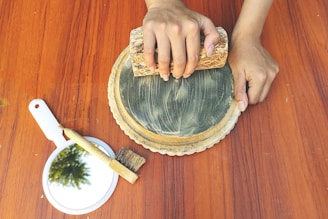 A pair of hands is holding a rectangular stone while grinding or smoothing a circular stone surface placed on a wooden table. Beside this setup, there is a handheld mirror with a reflection of greenery and a small brush.