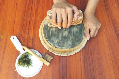 A pair of hands is holding a rectangular stone while grinding or smoothing a circular stone surface placed on a wooden table. Beside this setup, there is a handheld mirror with a reflection of greenery and a small brush.