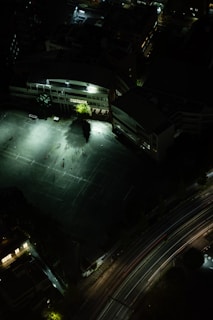 A night aerial view of an outdoor sports field adjacent to a large building. The field is illuminated by bright artificial lights, with some people visible as shadows on the ground. The surrounding area is mostly dark, creating a stark contrast. A curved road with light trails from vehicles runs parallel to the field.