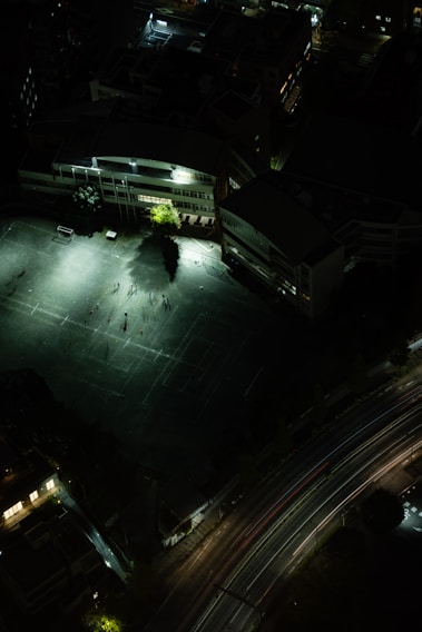 A night aerial view of an outdoor sports field adjacent to a large building. The field is illuminated by bright artificial lights, with some people visible as shadows on the ground. The surrounding area is mostly dark, creating a stark contrast. A curved road with light trails from vehicles runs parallel to the field.