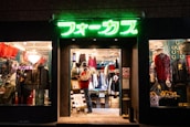 A brightly lit vintage clothing store with a neon green sign above the entrance. The interior showcases various garments, including jackets and shirts, displayed on racks. A mannequin dressed in winter attire stands near the entrance with sale signs visible. The window displays feature eclectic decorations and apparel.