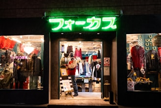 A brightly lit vintage clothing store with a neon green sign above the entrance. The interior showcases various garments, including jackets and shirts, displayed on racks. A mannequin dressed in winter attire stands near the entrance with sale signs visible. The window displays feature eclectic decorations and apparel.