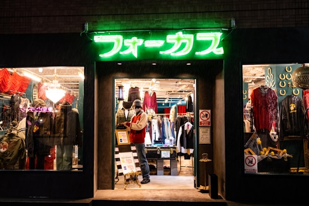 A brightly lit vintage clothing store with a neon green sign above the entrance. The interior showcases various garments, including jackets and shirts, displayed on racks. A mannequin dressed in winter attire stands near the entrance with sale signs visible. The window displays feature eclectic decorations and apparel.
