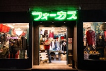 A brightly lit vintage clothing store with a neon green sign above the entrance. The interior showcases various garments, including jackets and shirts, displayed on racks. A mannequin dressed in winter attire stands near the entrance with sale signs visible. The window displays feature eclectic decorations and apparel.
