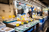 A bustling fish market with numerous stalls displaying a variety of fresh seafood on ice, including fish, shrimp, and other marine products. Each stall is equipped with bright signs indicating prices. Several vendors, wearing aprons and masks, are actively engaging with the produce and customers in a well-lit, busy environment.