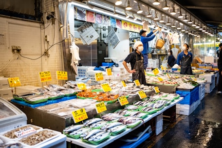 A bustling fish market with numerous stalls displaying a variety of fresh seafood on ice, including fish, shrimp, and other marine products. Each stall is equipped with bright signs indicating prices. Several vendors, wearing aprons and masks, are actively engaging with the produce and customers in a well-lit, busy environment.