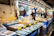 A bustling fish market with numerous stalls displaying a variety of fresh seafood on ice, including fish, shrimp, and other marine products. Each stall is equipped with bright signs indicating prices. Several vendors, wearing aprons and masks, are actively engaging with the produce and customers in a well-lit, busy environment.
