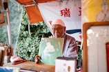 An elderly man wearing glasses and a white headscarf sits under a canopy, holding up a piece of art featuring a white coiled shape against a green background. The setting appears to be outdoors, with leafy greenery visible in the background. Various art supplies, including brushes, are arranged on the table in front of him. A banner with red and blue text hangs behind the man.