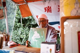 An elderly man wearing glasses and a white headscarf sits under a canopy, holding up a piece of art featuring a white coiled shape against a green background. The setting appears to be outdoors, with leafy greenery visible in the background. Various art supplies, including brushes, are arranged on the table in front of him. A banner with red and blue text hangs behind the man.
