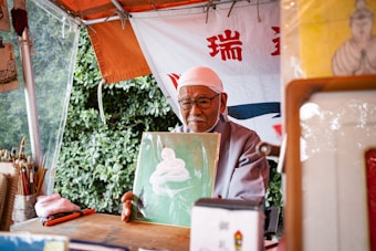 An elderly man wearing glasses and a white headscarf sits under a canopy, holding up a piece of art featuring a white coiled shape against a green background. The setting appears to be outdoors, with leafy greenery visible in the background. Various art supplies, including brushes, are arranged on the table in front of him. A banner with red and blue text hangs behind the man.