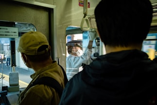 Inside a train, two passengers stand near the door, one wearing a cap and the other in dark clothing. A conductor is visible outside yelling commands, wearing a uniform and mask, while holding onto the train. Reflections on the glass hint at the bustling station environment.