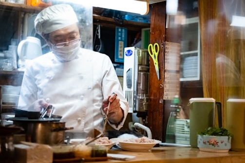 A chef wearing a white uniform, face mask, and chef's hat is preparing food in a kitchen. There are plates of food on the counter, a pot on the stove, and various kitchen utensils and appliances in the background. A pair of yellow scissors is hanging on the wooden wall, and there are some containers and a floral-patterned bowl with greenery next to the chef.