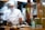 A chef wearing a white uniform, face mask, and chef's hat is preparing food in a kitchen. There are plates of food on the counter, a pot on the stove, and various kitchen utensils and appliances in the background. A pair of yellow scissors is hanging on the wooden wall, and there are some containers and a floral-patterned bowl with greenery next to the chef.