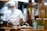 A chef wearing a white uniform, face mask, and chef's hat is preparing food in a kitchen. There are plates of food on the counter, a pot on the stove, and various kitchen utensils and appliances in the background. A pair of yellow scissors is hanging on the wooden wall, and there are some containers and a floral-patterned bowl with greenery next to the chef.