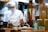 A chef wearing a white uniform, face mask, and chef's hat is preparing food in a kitchen. There are plates of food on the counter, a pot on the stove, and various kitchen utensils and appliances in the background. A pair of yellow scissors is hanging on the wooden wall, and there are some containers and a floral-patterned bowl with greenery next to the chef.