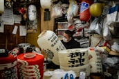 A person wearing a mask works inside a cozy, cluttered workshop filled with traditional Japanese lanterns. There are various lanterns hanging and stacked around, with some displaying intricate designs and calligraphy. The workshop has wooden walls and is filled with bags, tools, and materials for crafting.