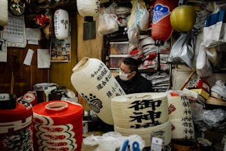 A person wearing a mask works inside a cozy, cluttered workshop filled with traditional Japanese lanterns. There are various lanterns hanging and stacked around, with some displaying intricate designs and calligraphy. The workshop has wooden walls and is filled with bags, tools, and materials for crafting.
