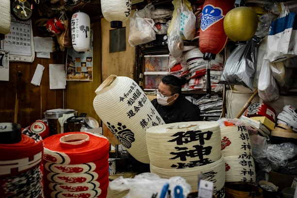 A person wearing a mask works inside a cozy, cluttered workshop filled with traditional Japanese lanterns. There are various lanterns hanging and stacked around, with some displaying intricate designs and calligraphy. The workshop has wooden walls and is filled with bags, tools, and materials for crafting.