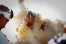 A person dressed in traditional attire holds a ceremonial object amidst a cloud of smoke, suggesting a ritual or cultural event. The focus is on the individual's contemplative expression, with a background blurred by smoke.