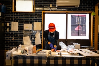 A person wearing a red cap and blue apron is working at a counter. The workspace has various kitchen items, including bottles, containers, and a large knife. The background features dark blue tile walls, a window with a poster, and visible paperwork.