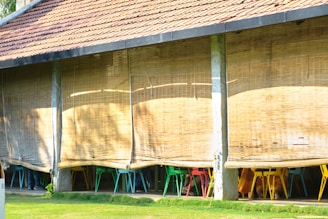 An outdoor seating area with bamboo blinds partially rolled down, revealing colorful chairs underneath. The structure has a tiled roof and is supported by vertical stone pillars. Sunlight casts shadows on the bamboo blinds, creating a warm and inviting ambiance.