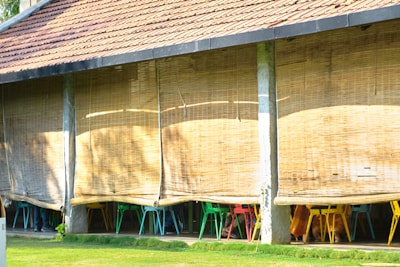 An outdoor seating area with bamboo blinds partially rolled down, revealing colorful chairs underneath. The structure has a tiled roof and is supported by vertical stone pillars. Sunlight casts shadows on the bamboo blinds, creating a warm and inviting ambiance.